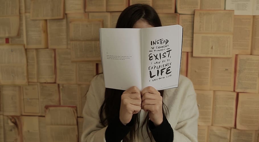 woman covering her face with book, woman holding a book with Instead Exist Life text near wall HD wallpaper