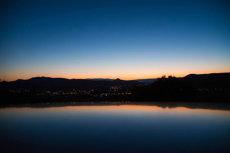 calm lake waters reflecting city and trees and clear sky, silhouette of buildings with lights view from body of water HD wallpaper