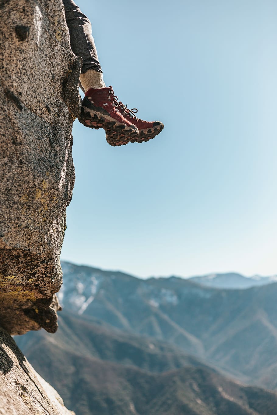 person wearing pair of red-and-white hiking shoes sitting on rocky cliff across mountains during daytime, person sitting on top of mountain at daytime HD wallpaper