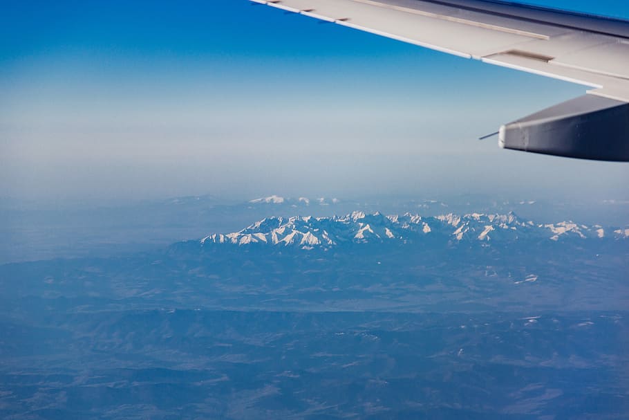 white airplane wing above a snow mountains, airplane above mountain under white clouds and blue sky