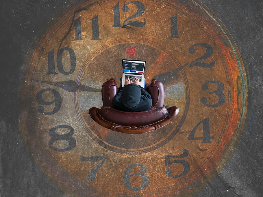 brown analog clock, top view photography of man sitting on brown leather sofa chair while using gray and black laptop computer
