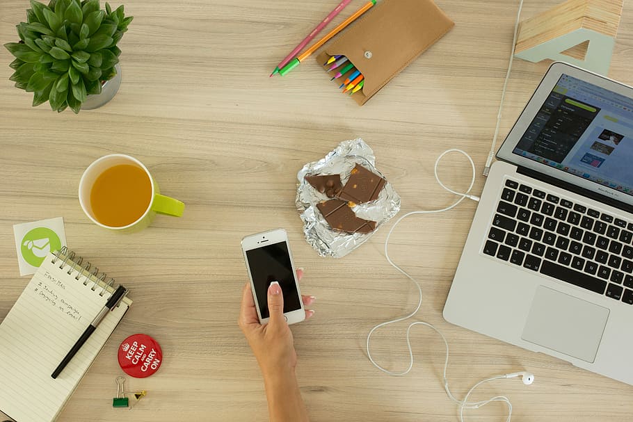 photo of person holding smartphone surrounded with laptop, chocolates, pens, notes, and cup with coffee HD wallpaper