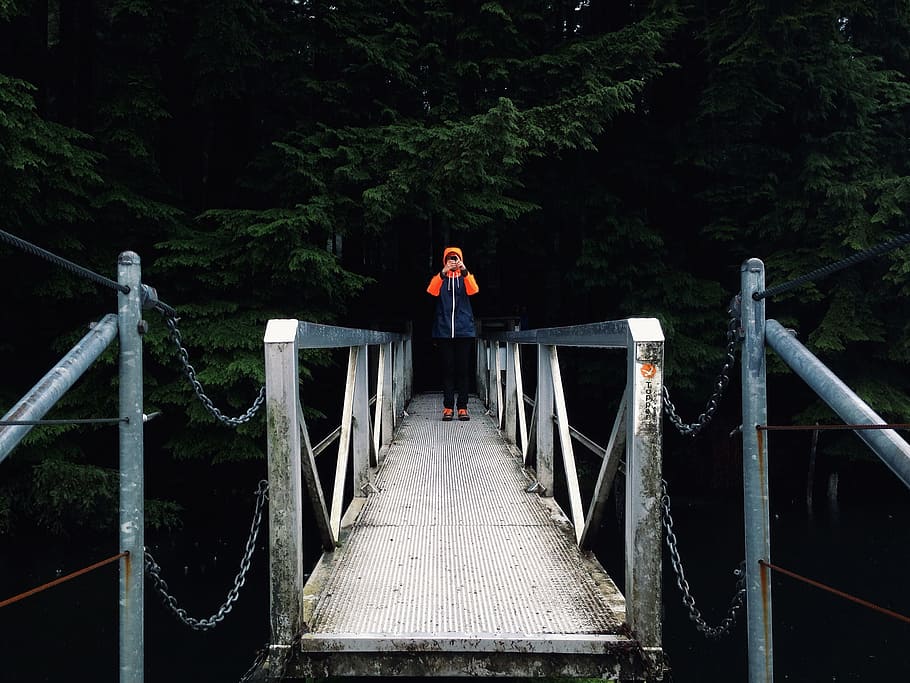 person standing on a bridge during day time, person standing on gray footbridge leading to forest HD wallpaper