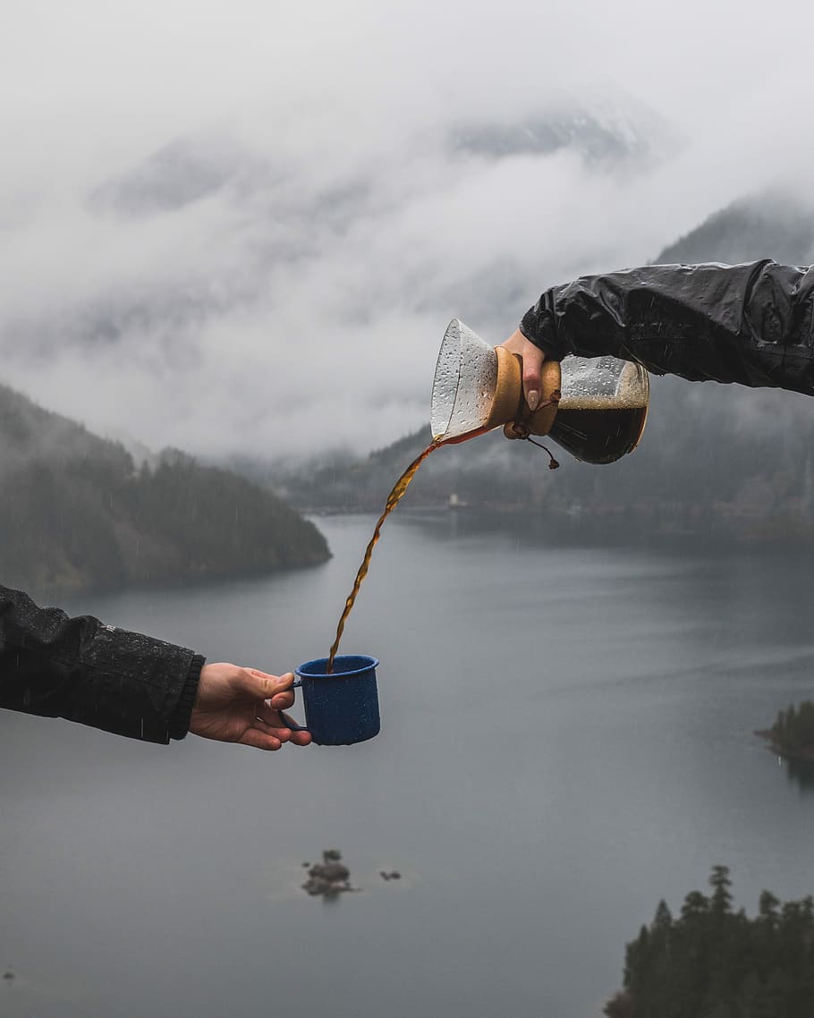 person pouring coffee to the another person's cup near body of water, person pouring coffee into blue ceramic mug from glass pitcher