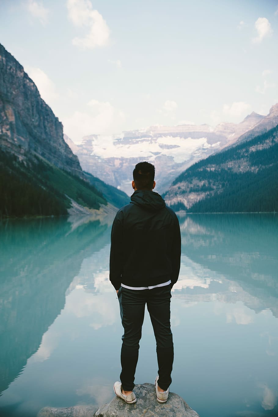 man standing in the middle rock facing lake, man in black hoodie and black pants standing in front of body of water and mountain during daytime HD wallpaper