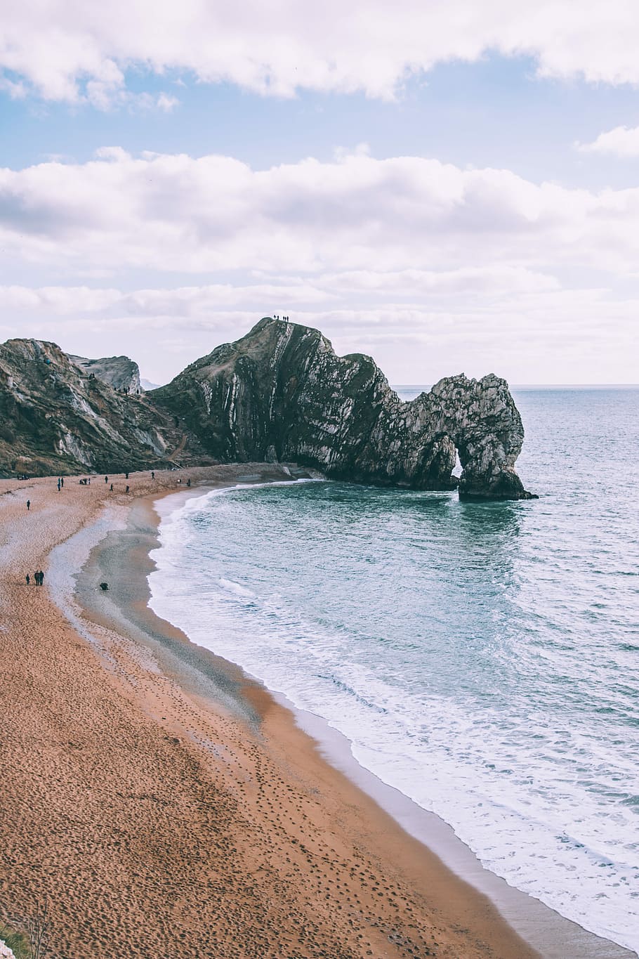 Cliffs on sand beach, people on seashore near gray rock formation HD wallpaper