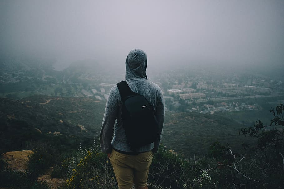man in gray hoodie with black backpack looking at the city from mountain peek, man in gray pullover hoodie on hill top during fog