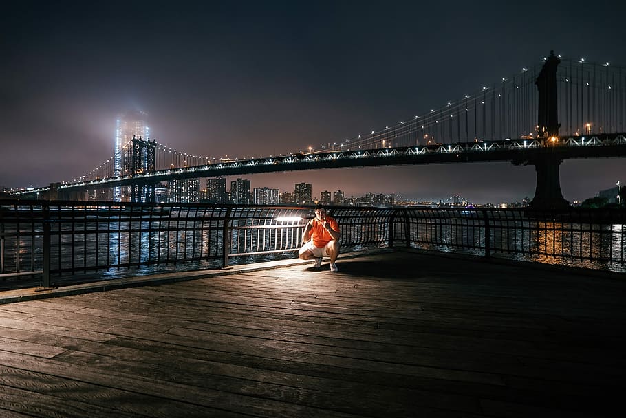 man sitting on dock in front bridge, photography man in orange top under full-suspension bridge HD wallpaper