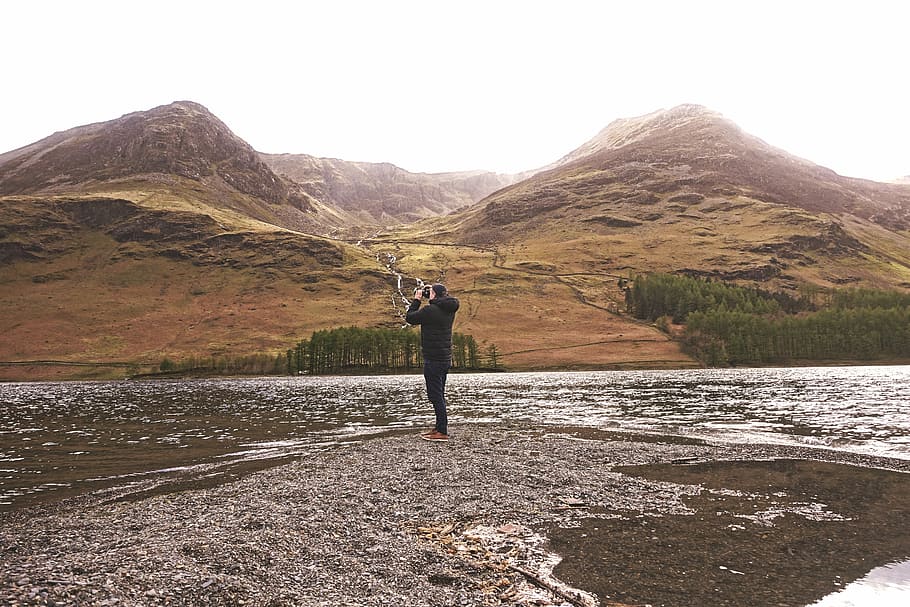 person taking photo of mountain, man standing near body of water during daytime HD wallpaper