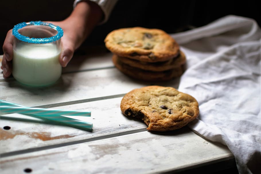 brown cookies on white wooden surface, selective focus photography of person holding glass of milk beside cookie on tray HD wallpaper