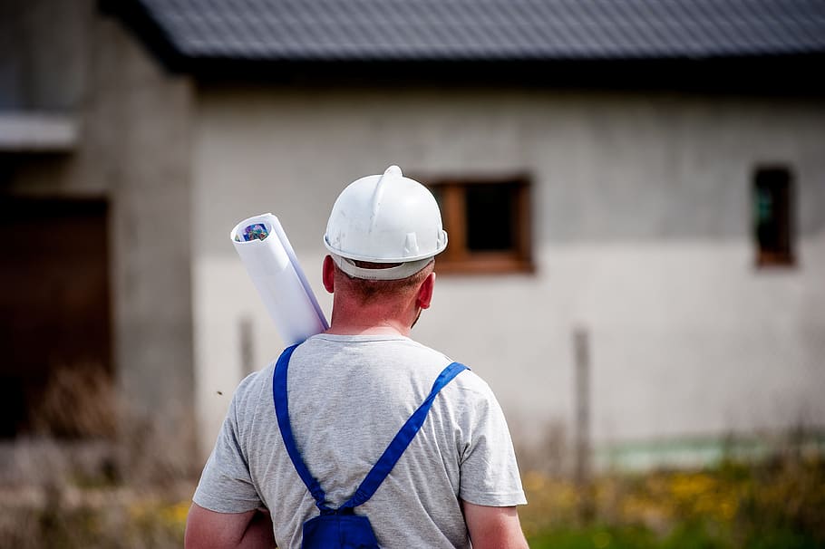man wearing white hard hat, gray shirt and blue rompers carrying blueprints facing gray concrete building HD wallpaper