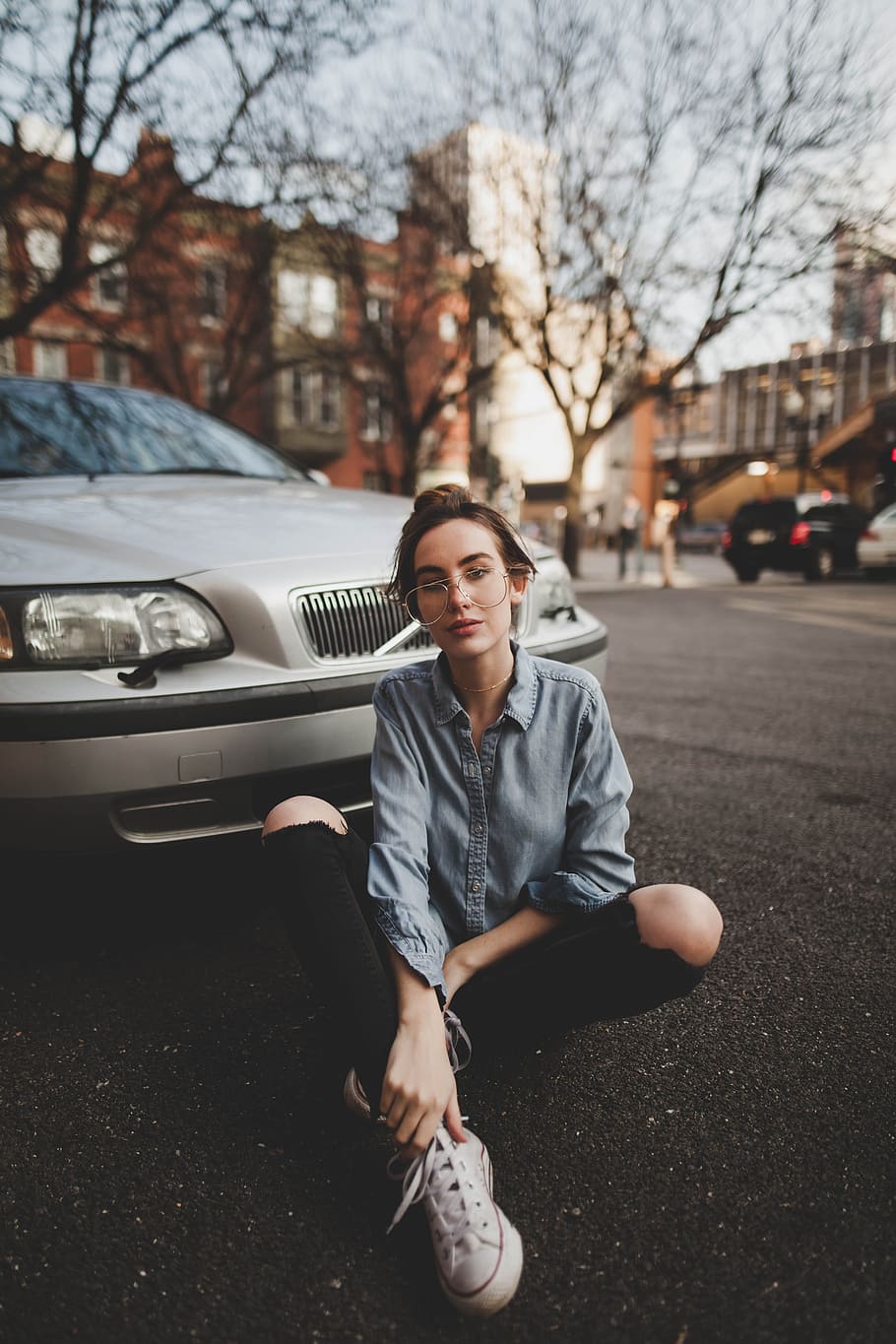 selective focus photography of woman wearing blue chambray shirt, black distressed denim skinny jeans, and pair of white high-top sneakers sitting on road in front of silver Volvo S60, woman in grey dress shirt sitting on ground