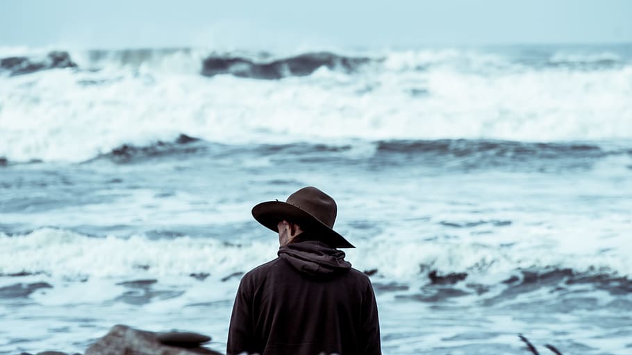 man wearing brown hat standing in front of ocean, man standing while wearing hat in front of body of water with wave during daytime HD wallpaper