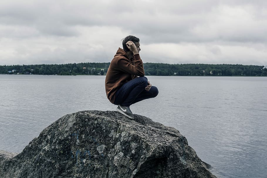 man wearing brown hoodie while standing on rock, man sitting on rock near body of water HD wallpaper