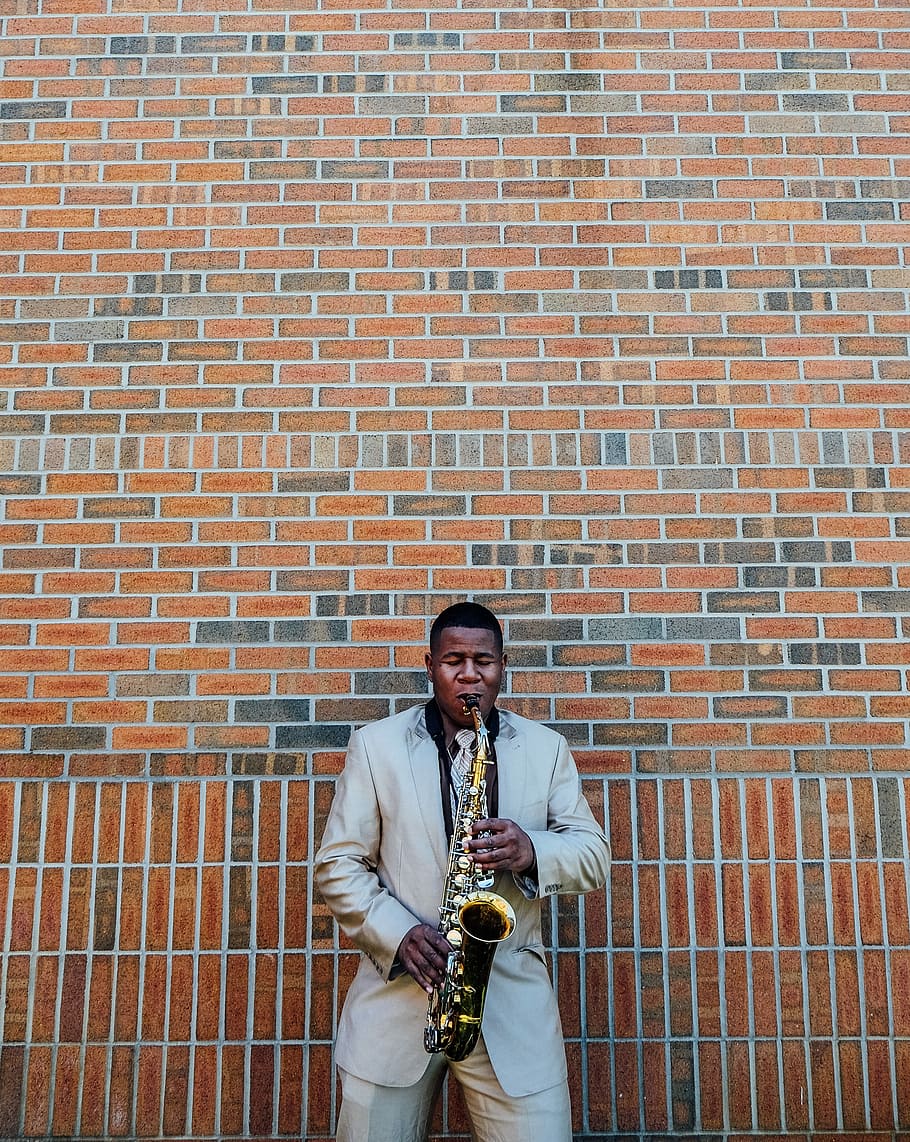 man playing saxophone near concrete brick, man wearing gray suit jacket playing saxophone in front of brown bricked building HD wallpaper