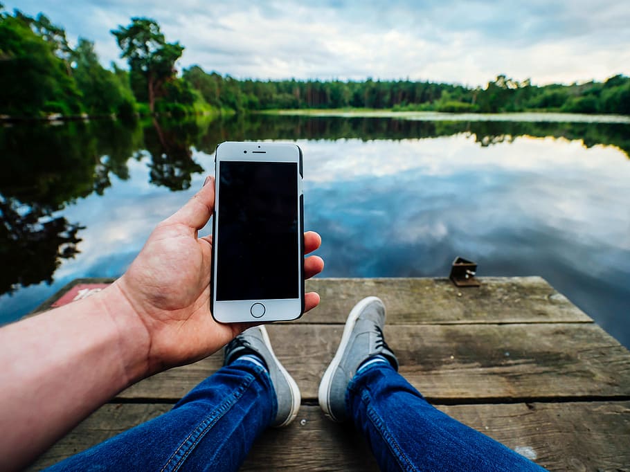 person sitting on river dock while holding iPhone near trees and under cumulus clouds HD wallpaper
