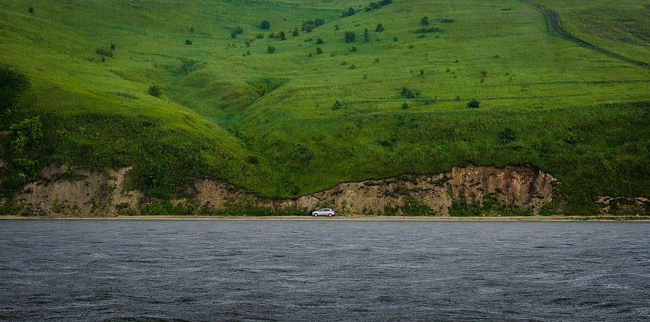 white car travelling on road in middle of hill and sea, silver vehicle on road near body of water and mountain HD wallpaper