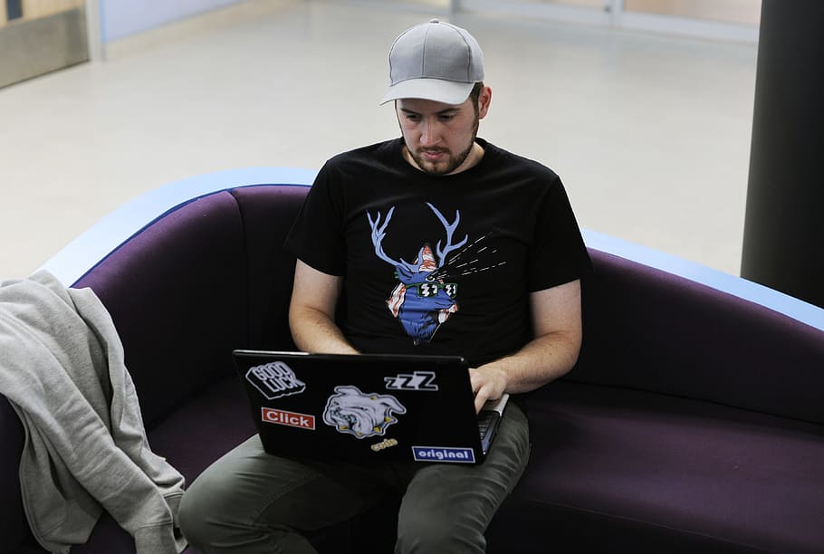 man wearing black crew neck shirt and grey baseball cap while using black laptop computer, person in black shirt sitting in purple sofa holding black laptop HD wallpaper