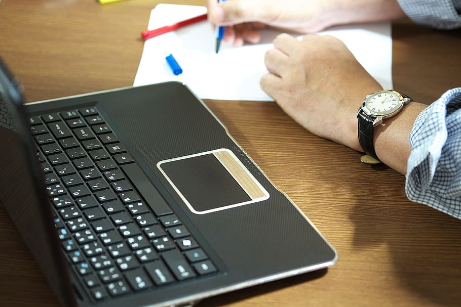 closeup photo of person holding blue pen writing on white paper beside black laptop computer HD wallpaper
