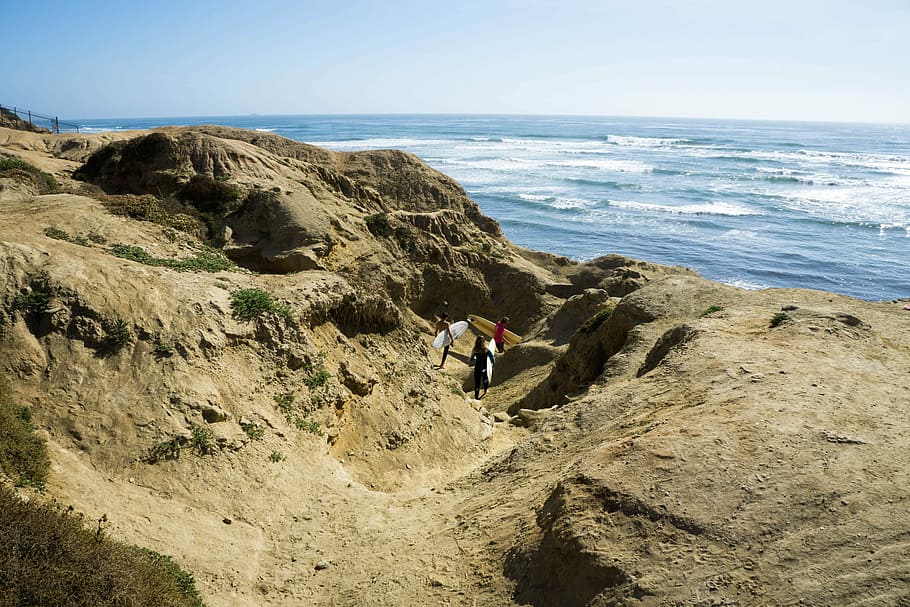 three persons carrying surfboards, three people carrying surfboards on brown rock formation under blue sky HD wallpaper