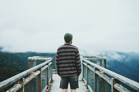 man standing on dock, man wearing brown-and-grey striped sweater, blue denim jeans, and green cap standing in the middle of metal bridge overlooking view full of trees under cloudy sky during daytime HD wallpaper