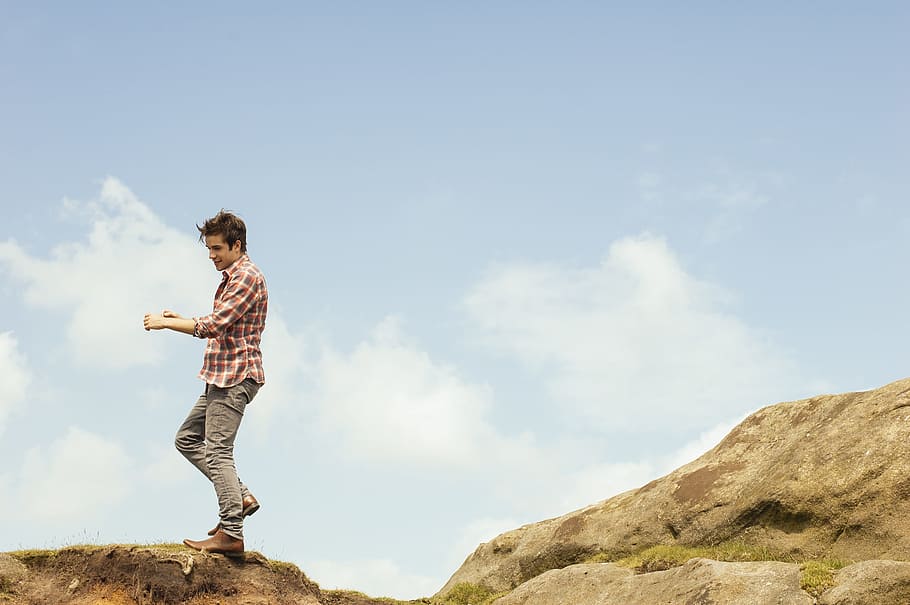 man in red plaid top standing on hilltop during daytime, man standing on top of rock