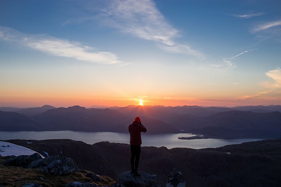 person standing on mountain peak facing body of water during sunrise HD wallpaper