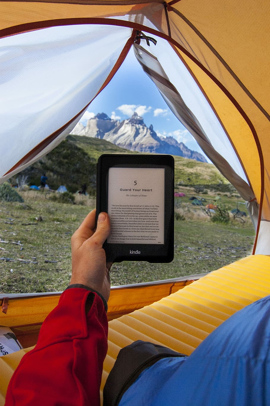 person holding black Amazon Kindle E-Book reader inside tent at daytime, person holding black e-book reader inside tent