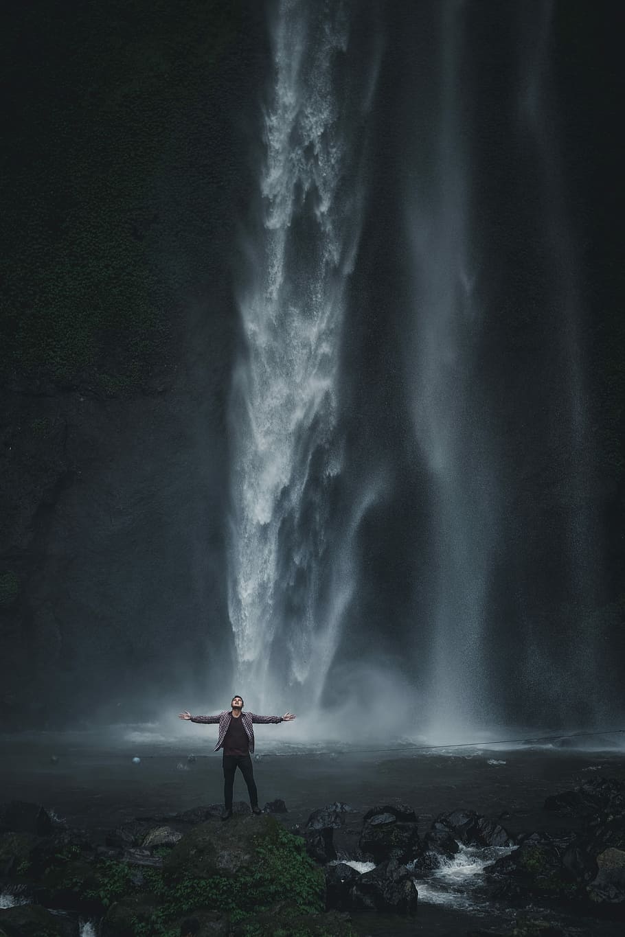 man in gray jacket standing near waterfalls, man standing on waterfalls while spreading his arms