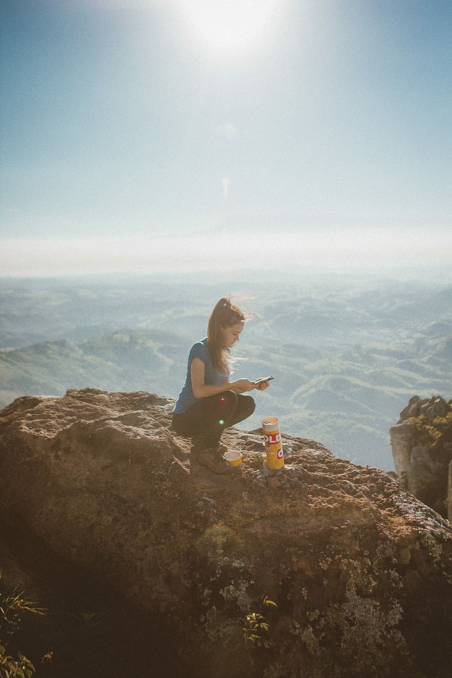 woman holding smartphone while sitting beside orange bottle on top of rock formation, woman using smartphone squating on brown rock in aerial photography