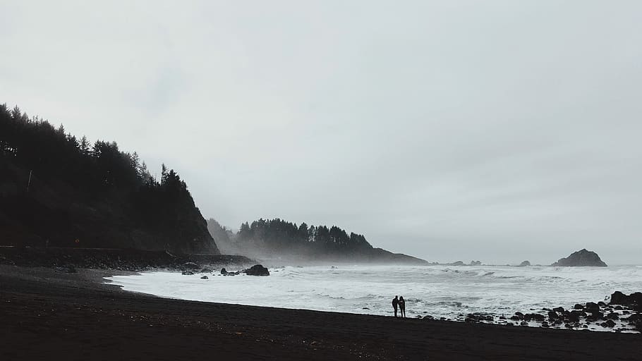 silhouette of 2 person standing near sea shore under gray sky HD wallpaper