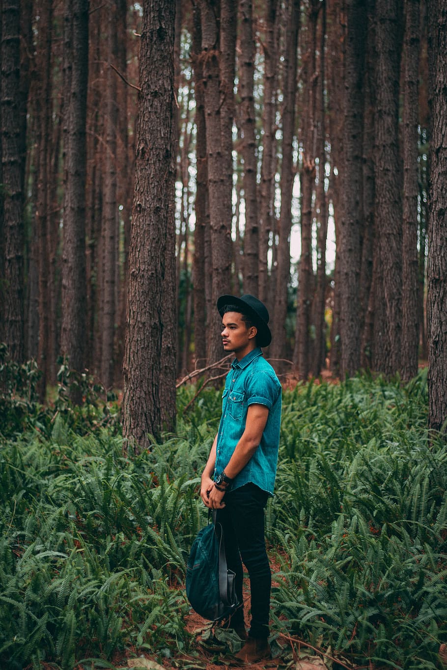 man standing in the middle of forest during daytime, man in green western shirt and black denim jeans with black fedora hat standing in forest holding green and black backpack photoshoot