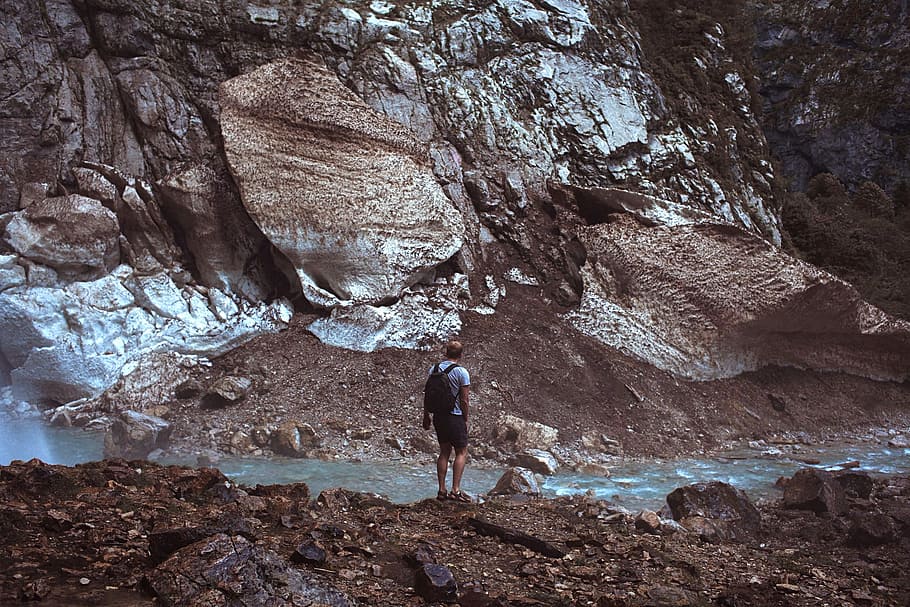 photography of man standing on rocks in front of river and mountain, man standing in front of body of water while carrying black backpack