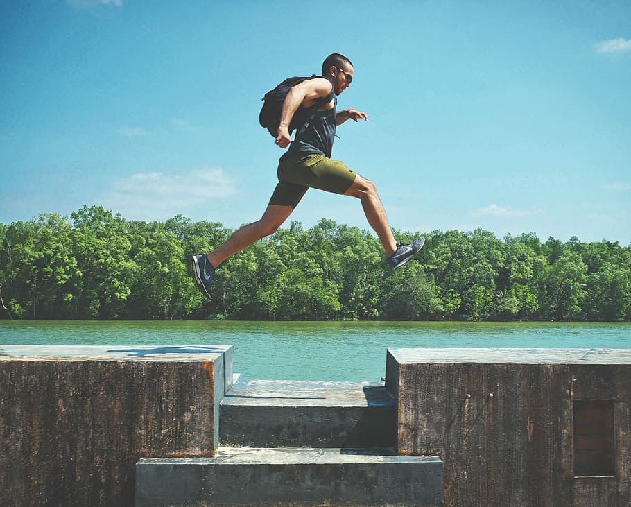 man leaping on concrete surface near body of water and forest at the distance during day, man jumping near river