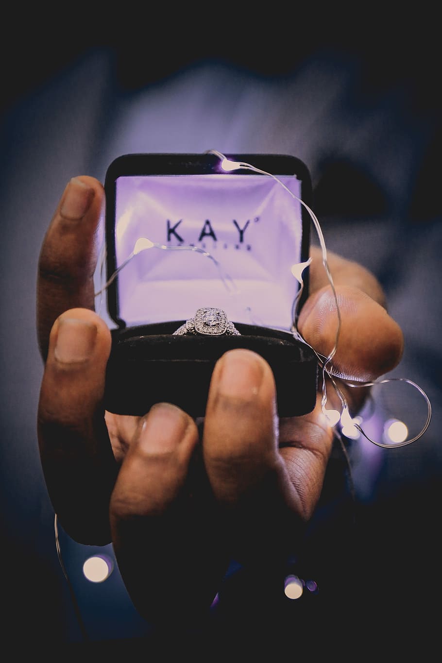 person holding silver-colored ring, person holding opened Kay Jeweler engagement ring box with string lights