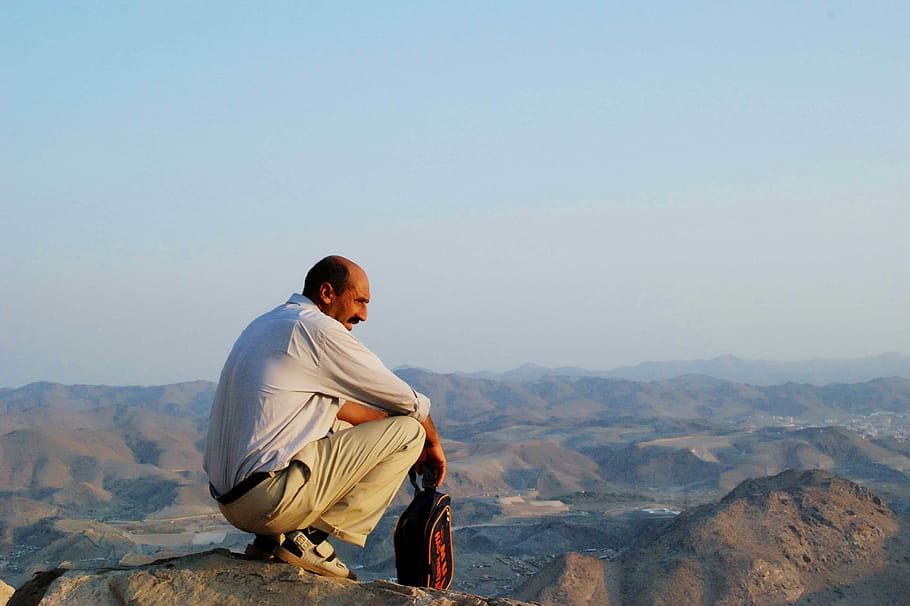 man wearing white dress shirt and khaki pants sitting on peak of mountain viewing hills at distance HD wallpaper