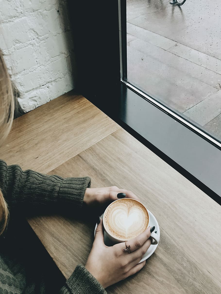 person holding cappuccino on brown wooden desk, person holding ceramic mug filled with coffee using both hands HD wallpaper
