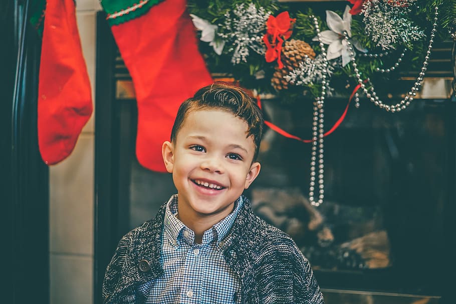 smiling boy in gray inner shirt and gray cardigan standing by fireplace with decors, portrait photo of boy standing near window and Christmas stockings HD wallpaper