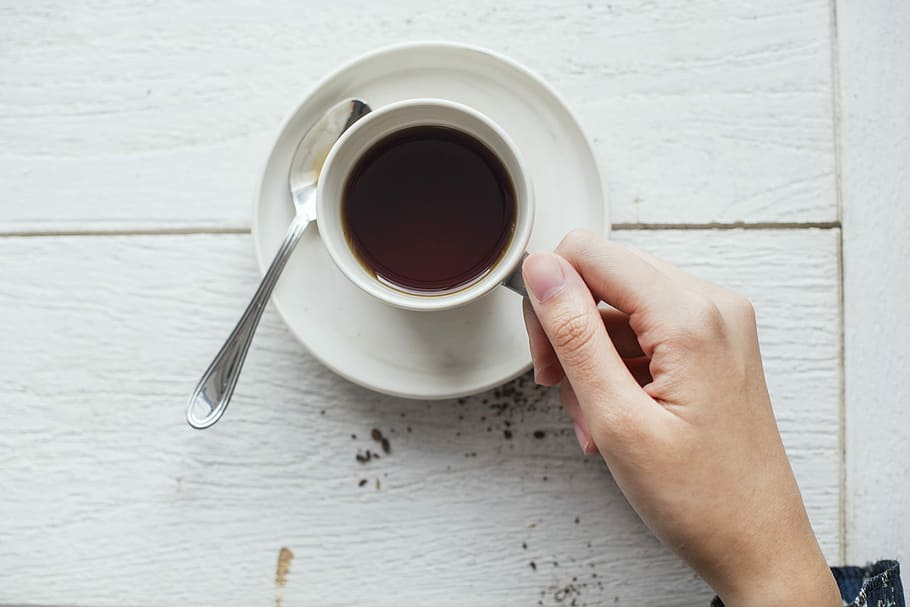 person holding cup of coffee on white saucer beside spoon, drink