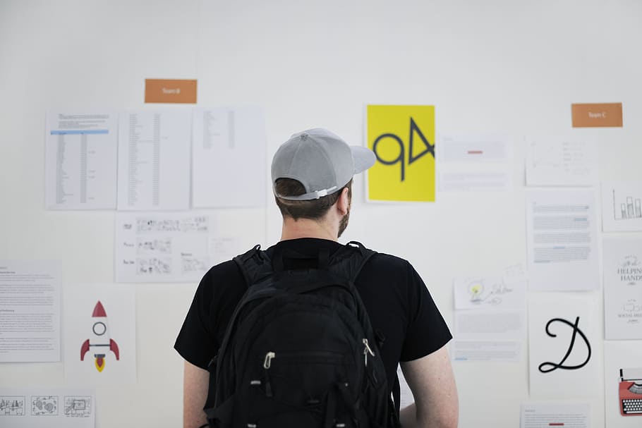 man wearing black shirt and gray cap in front of white board, man looking at bulletin board