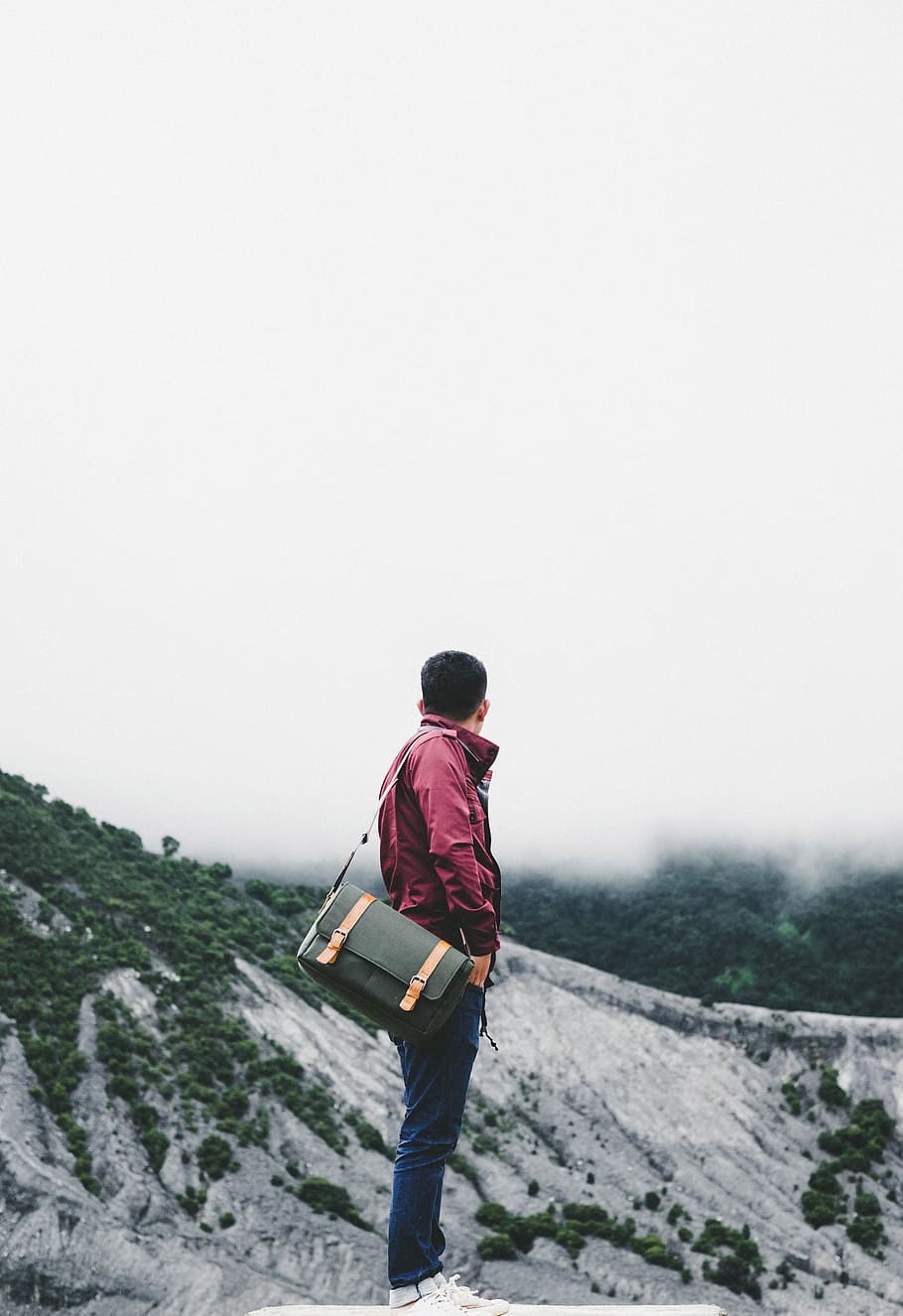man carrying black crossbody bag while standing on platform overlooking hill under white sky at daytime, landscape photograph of man facing mountain range under clear white sky HD wallpaper