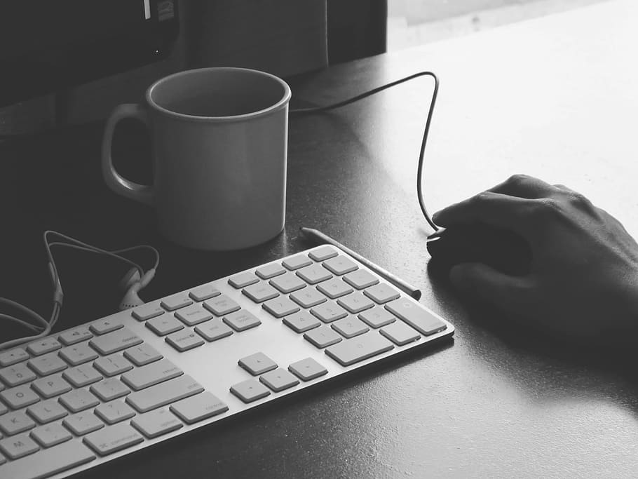 grayscale photography of person holding computer mouse near keyboard and mug, person holding mouse near keyboard