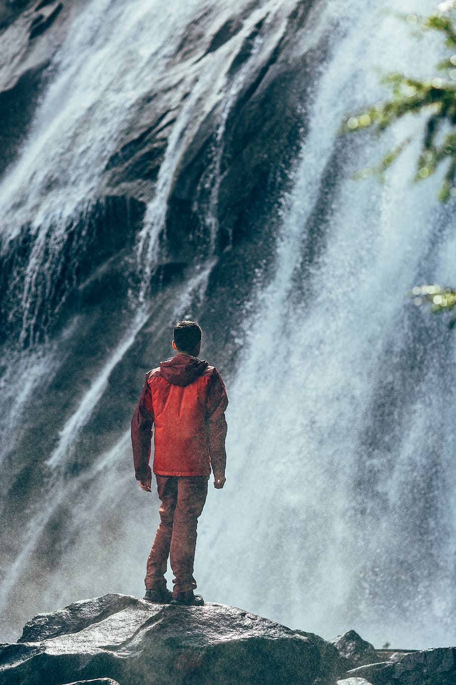 man wearing red hooded windbreaker jacket standing on gray rock while watching waterfalls at daytime, HD wallpaper
