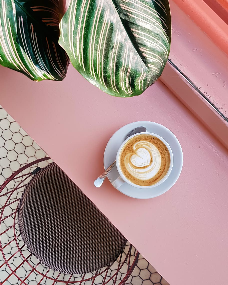 cup of coffee on saucer with teaspoon on pink tabletop, person taking picture of teacup on top of desk HD wallpaper