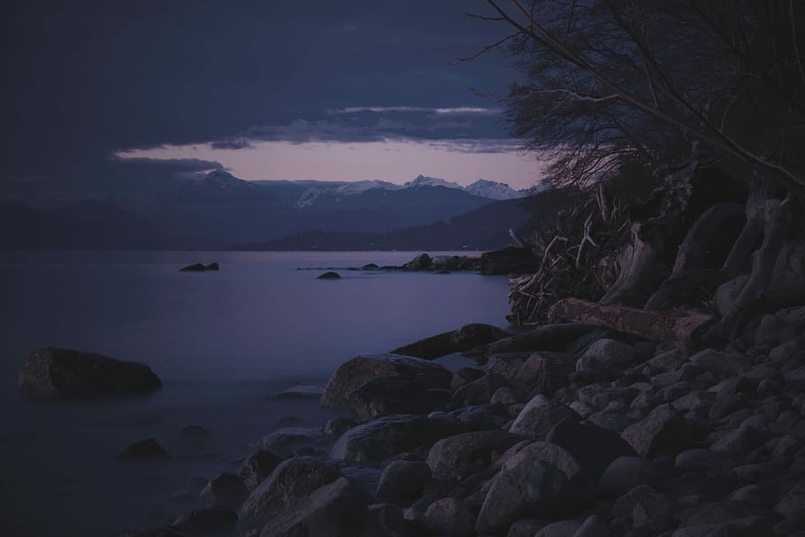 grey rock near body of water during dawn, rocks near calm body of water and mountains at daytime HD wallpaper