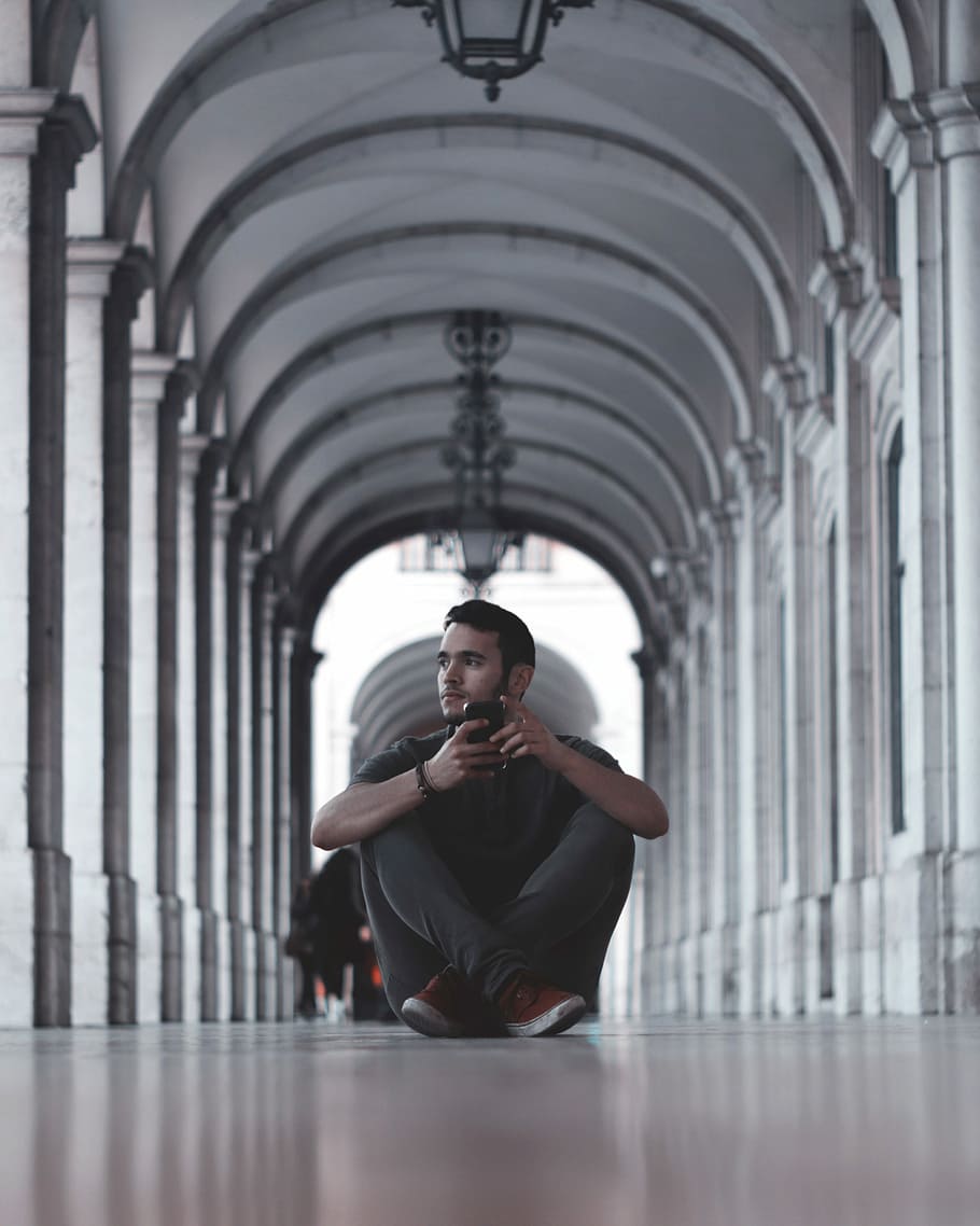 man wearing gray crew-neck and gray pants sitting on floor holding on smartphone, man holding black smartphone sitting near gray concrete pillars HD wallpaper