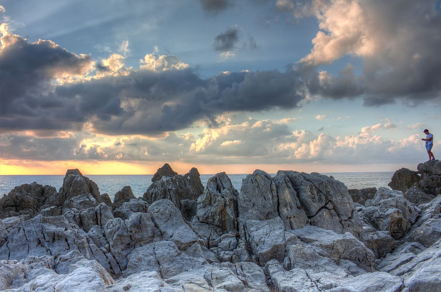 man standing on rock formation near ocean under gray clouds, person standing on top of gray large rocks HD wallpaper