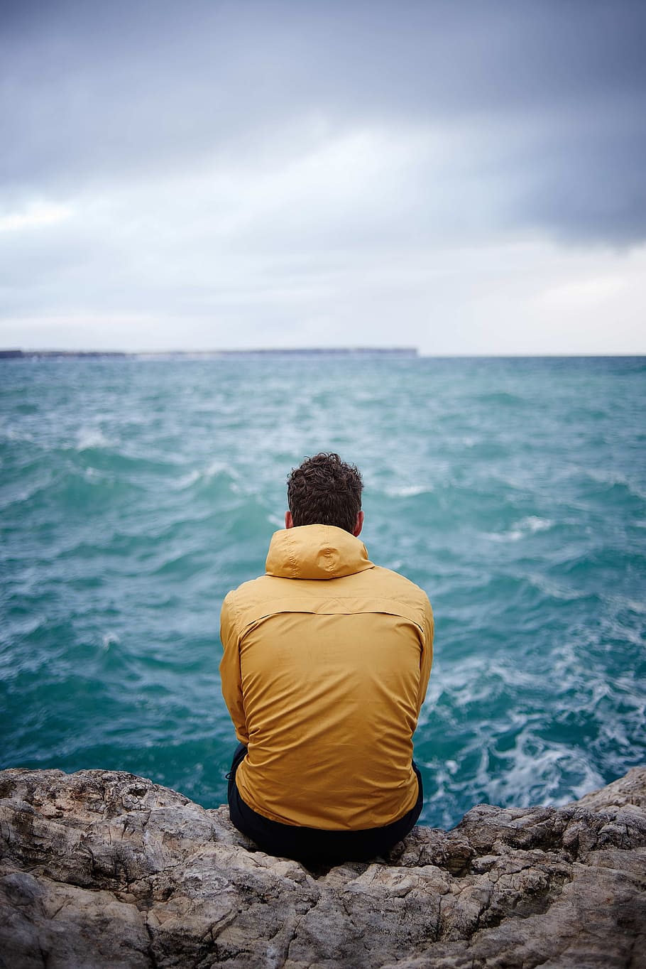man in yellow hooded jacket sitting on cliff in front of sea HD wallpaper