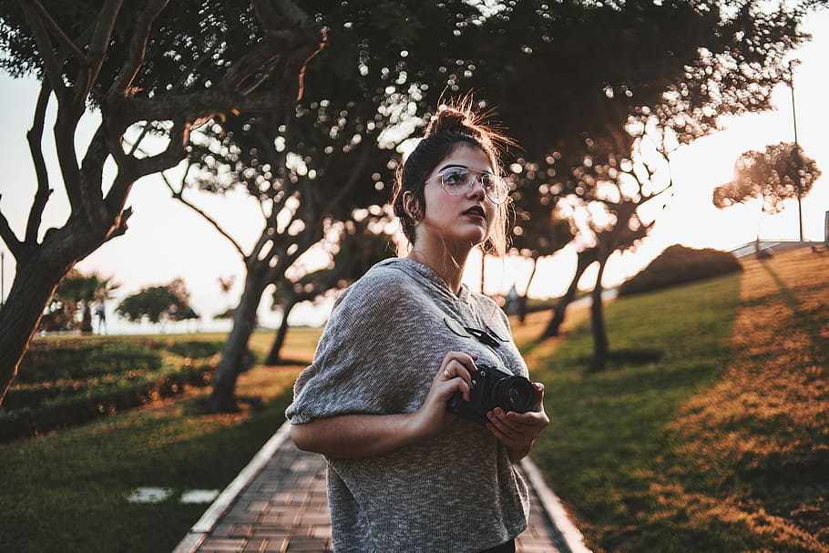 woman standing holding camera near tree, woman holding black camera near green leafed tree at daytime HD wallpaper