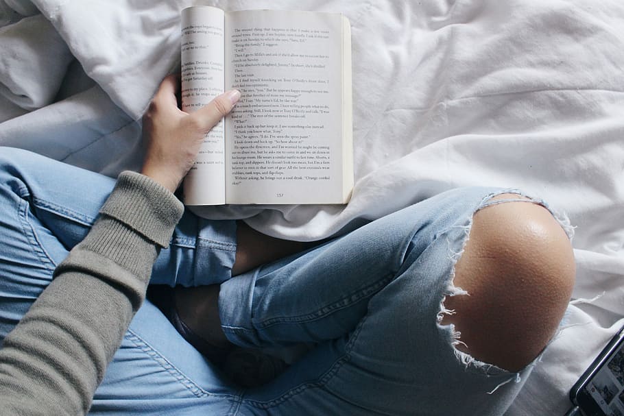 person reading book sitting with cross legs, person holding book on white textile HD wallpaper